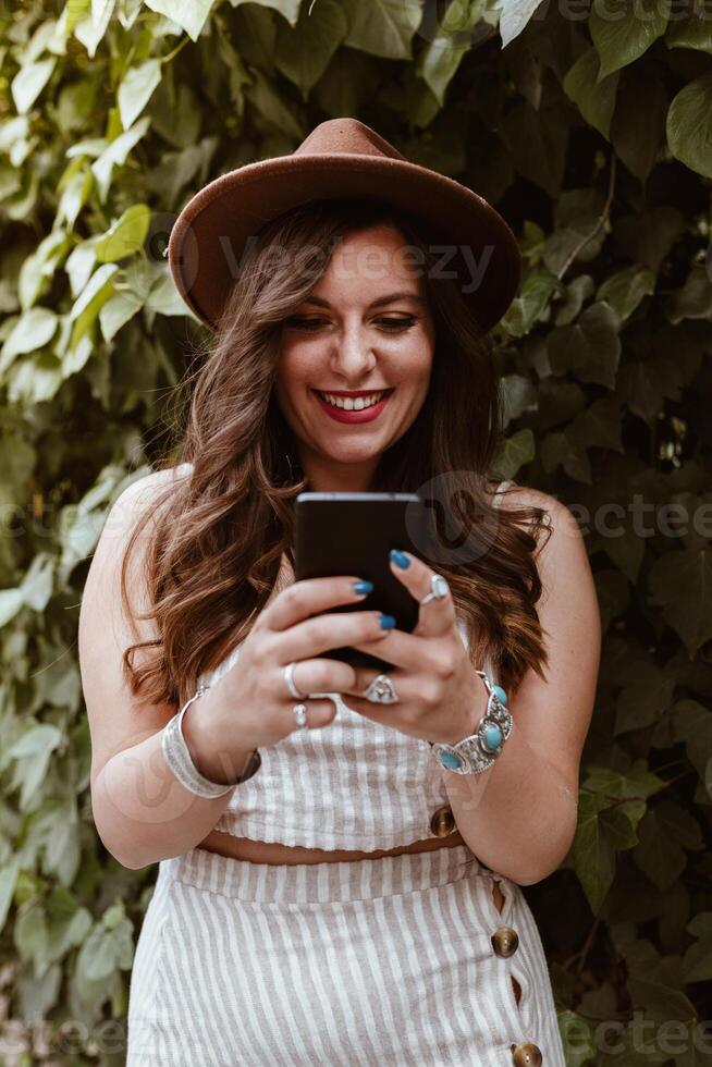 A woman in a hat and dress is smiling while using her phone photo