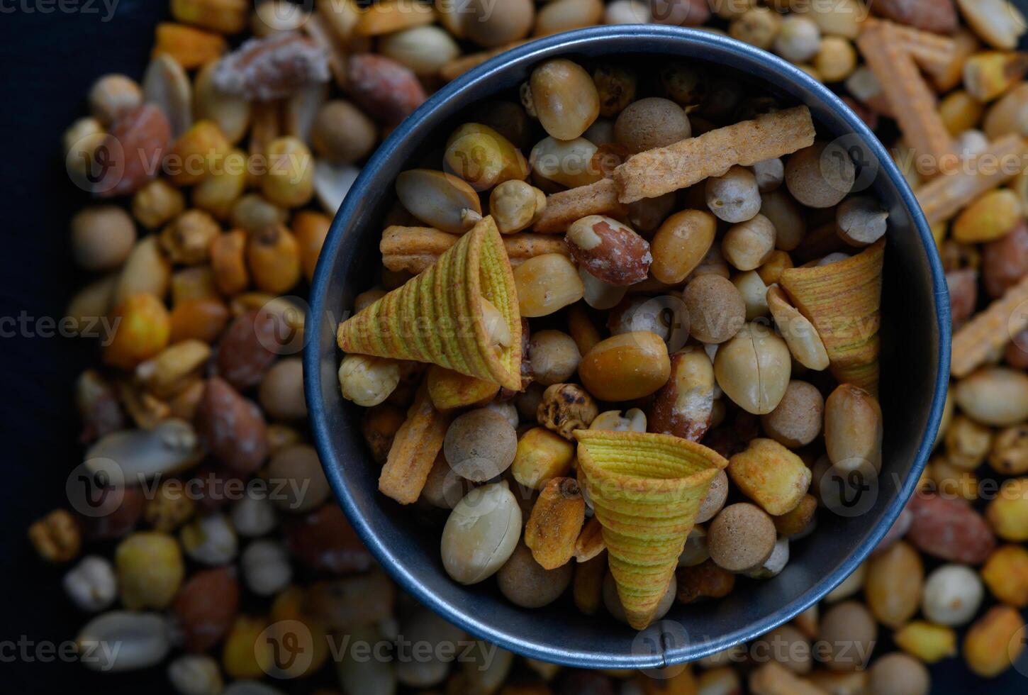 Colorful snack mix in a silver bowl surrounded by a variety of nuts and crunchy bites on a dark wooden surface photo