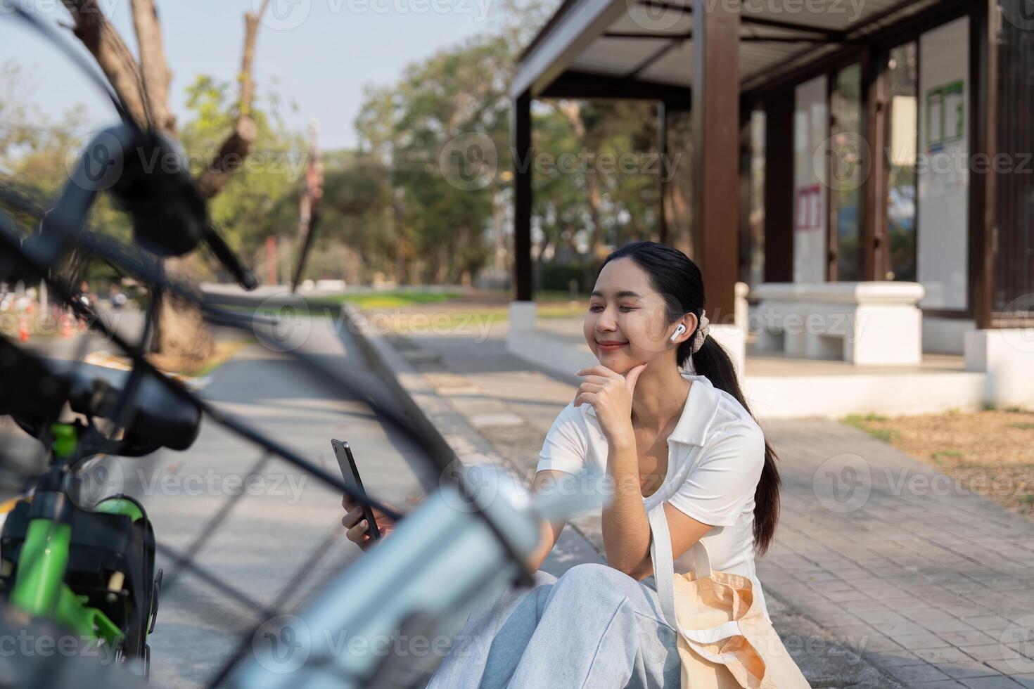 Relaxing with Technology and Nature. A woman comfortably seated beside her bike, enjoying music through earbuds while using her device outdoors. photo