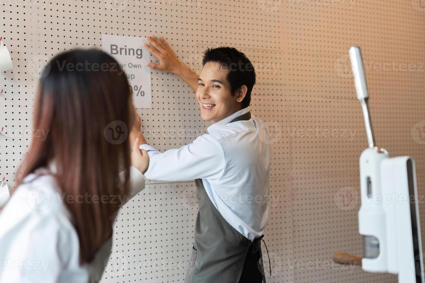 Eco-Friendly Promotion and cafe Community. A barista playfully engaging with her colleague while putting up a sign to encourage customers to bring their own cups for discounts. photo