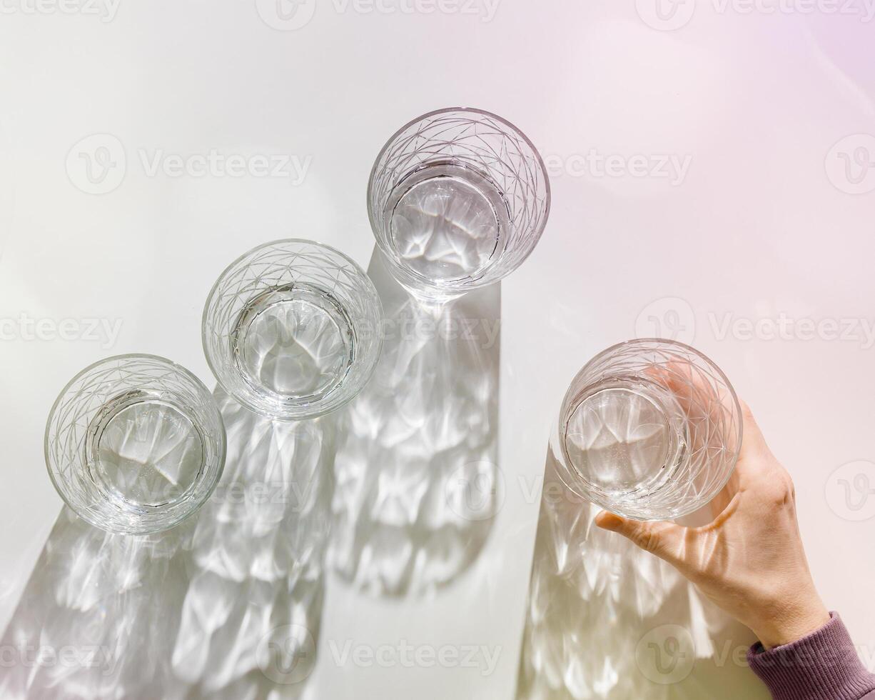 Transparent Water Glasses, hand holding one, shadows at sunlight on white, pastel color photo minimal style, aesthetic top view shiny cups sparkling at natural light, summer drink, composition