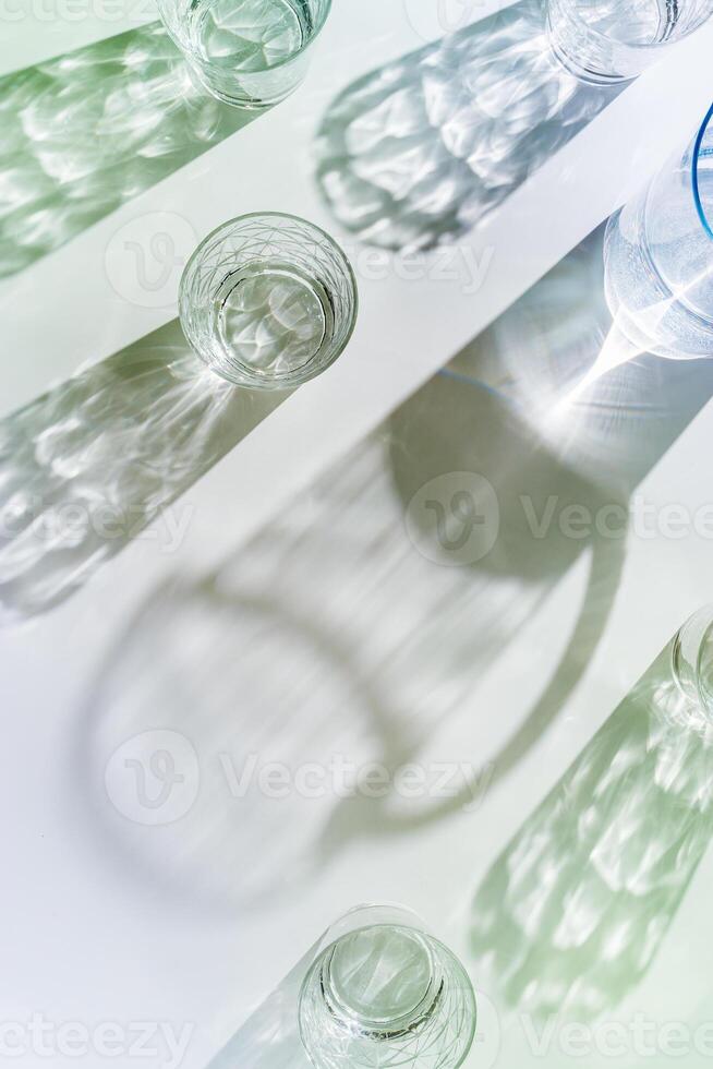 Transparent Water Glasses and water jug with shadows at sunlight on white, pastel color photo minimal style, aesthetic top view shiny cups sparkling at natural light, summer drink, composition