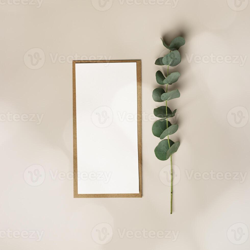 Blank paper card mock up and craft envelope, eucalyptus plant leaf on beige table. White empty letter or invitation, sunlight, beautiful soft shadow. Vertical lifestyle photo. Flat lay copyspace photo