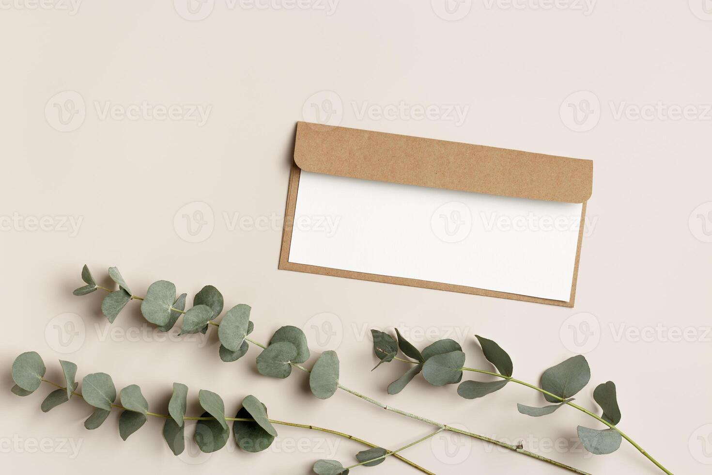 Blank paper card mock up and craft envelope with branches eucalyptus leaves on beige background. White empty invitation on table. Minimal lifestyle photo. Flat lay, top view, copy space. photo
