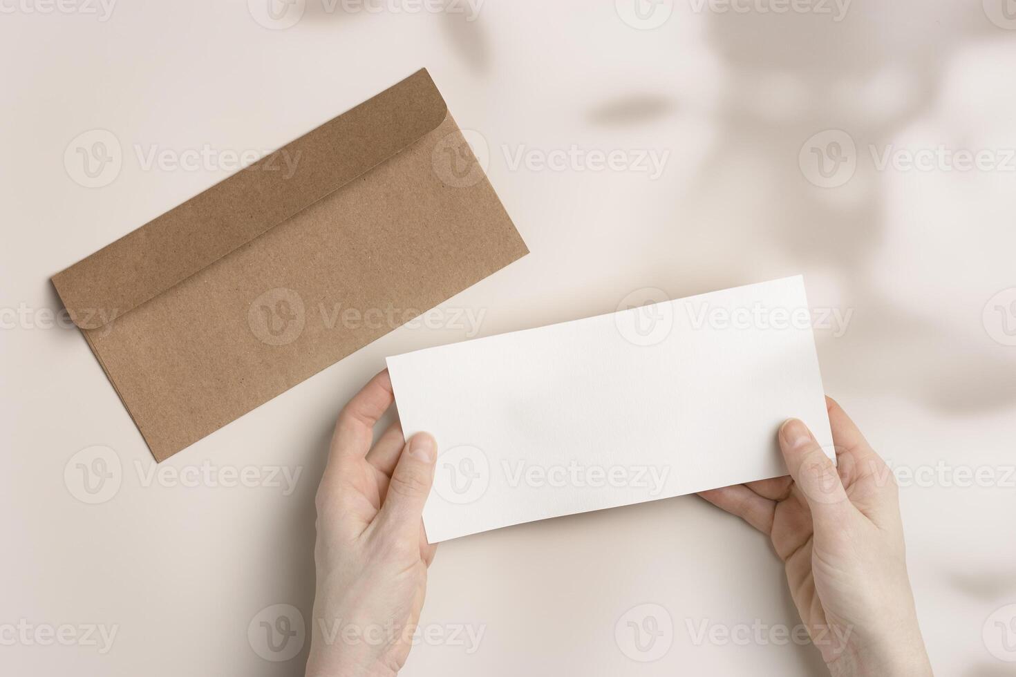 Blank paper card mock up in woman hands, craft envelope on beige background with shadow at sunlight. White empty invitation Female open paper letter. Minimal Flat lay, top view, copy space. photo