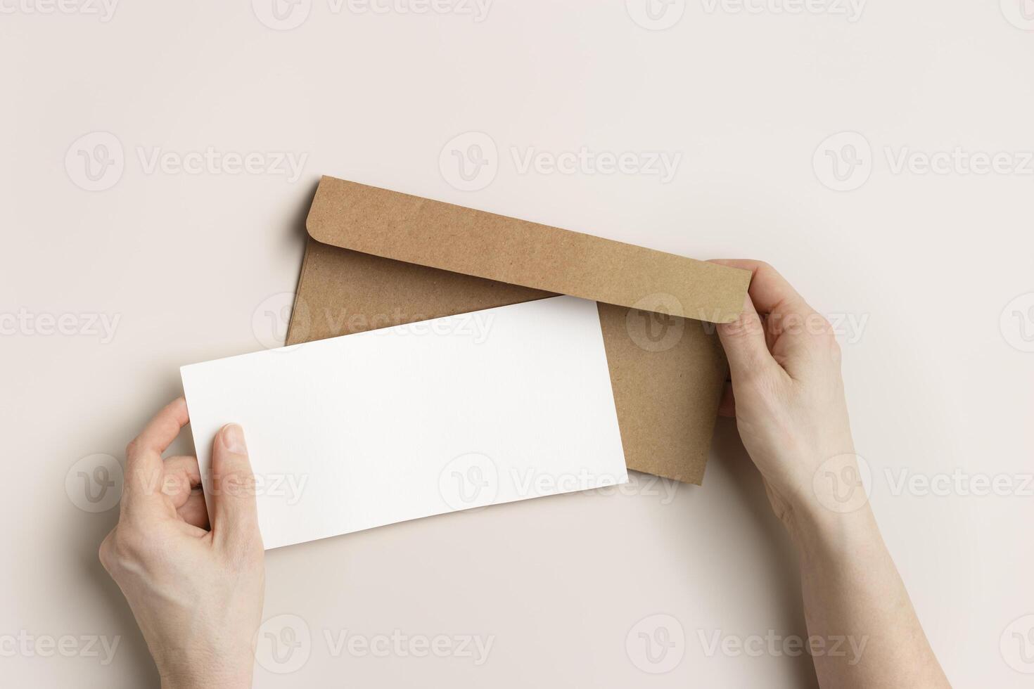 Blank paper card mock up in woman hands, craft envelope on beige background. White empty invitation on table. Female open paper letter. Minimal lifestyle photo. Flat lay, top view, copy space. photo
