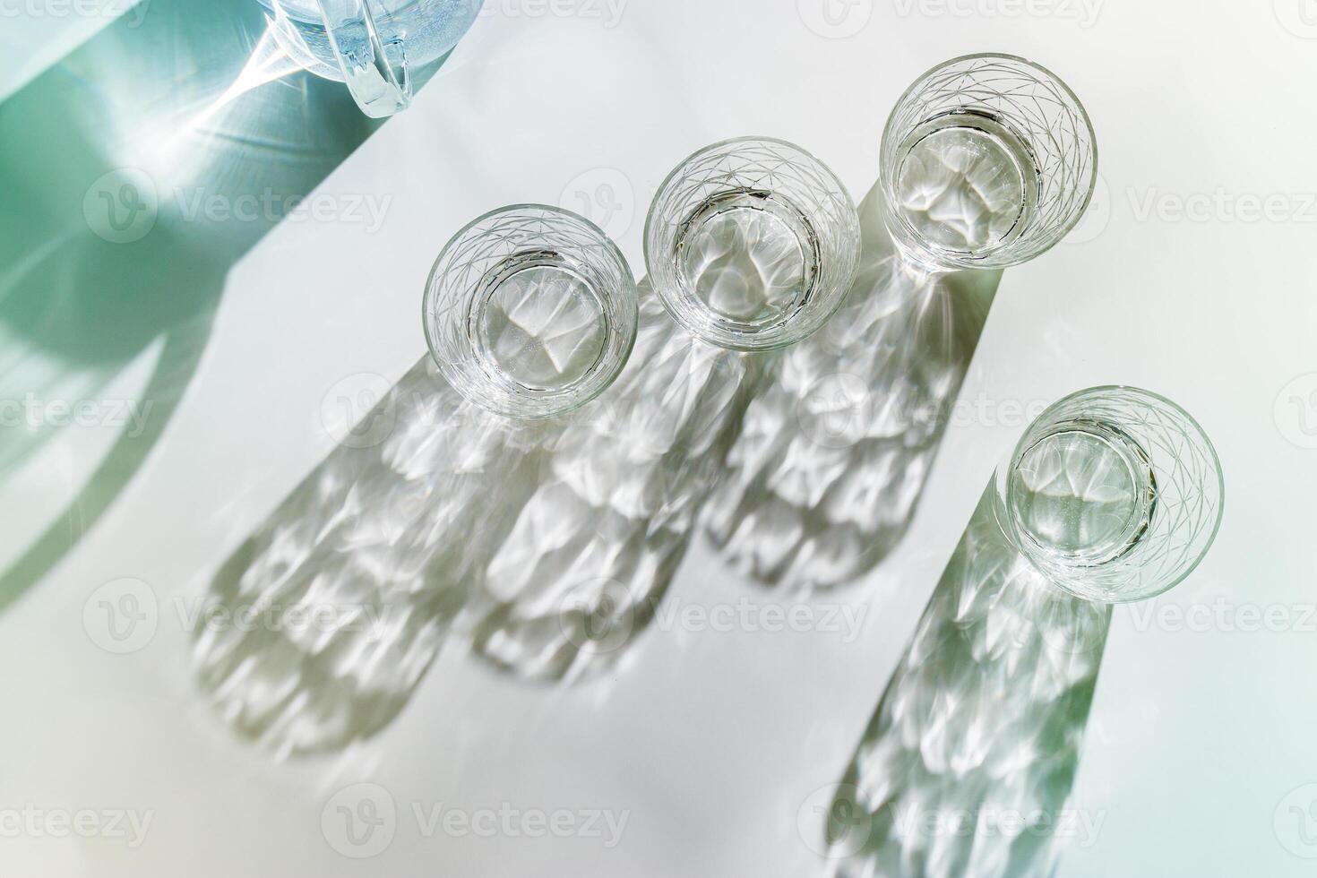 Transparent Water Glasses and water jug with shadows at sunlight on white, pastel color photo minimal style, aesthetic top view shiny cups sparkling at natural light, summer drink, composition.