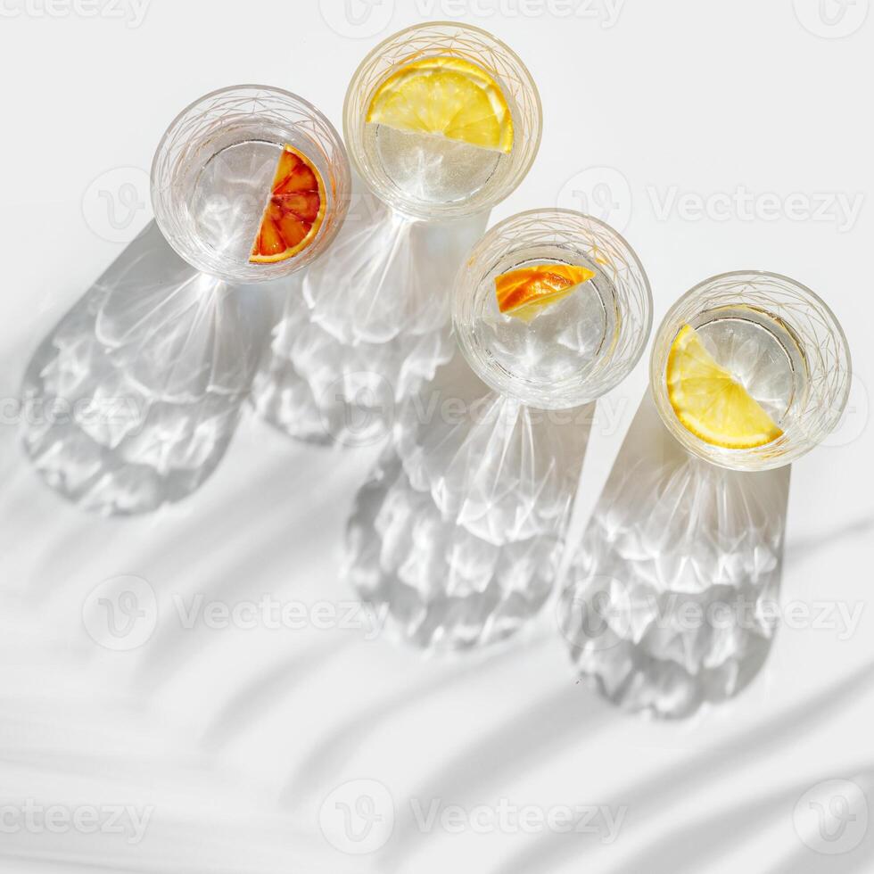 Crystal glasses with citrus slices in water on white background with palm leaf at sunlight, modern, minimalist composition with clear glass cups with water and citrus fruit, as lemon, orange photo
