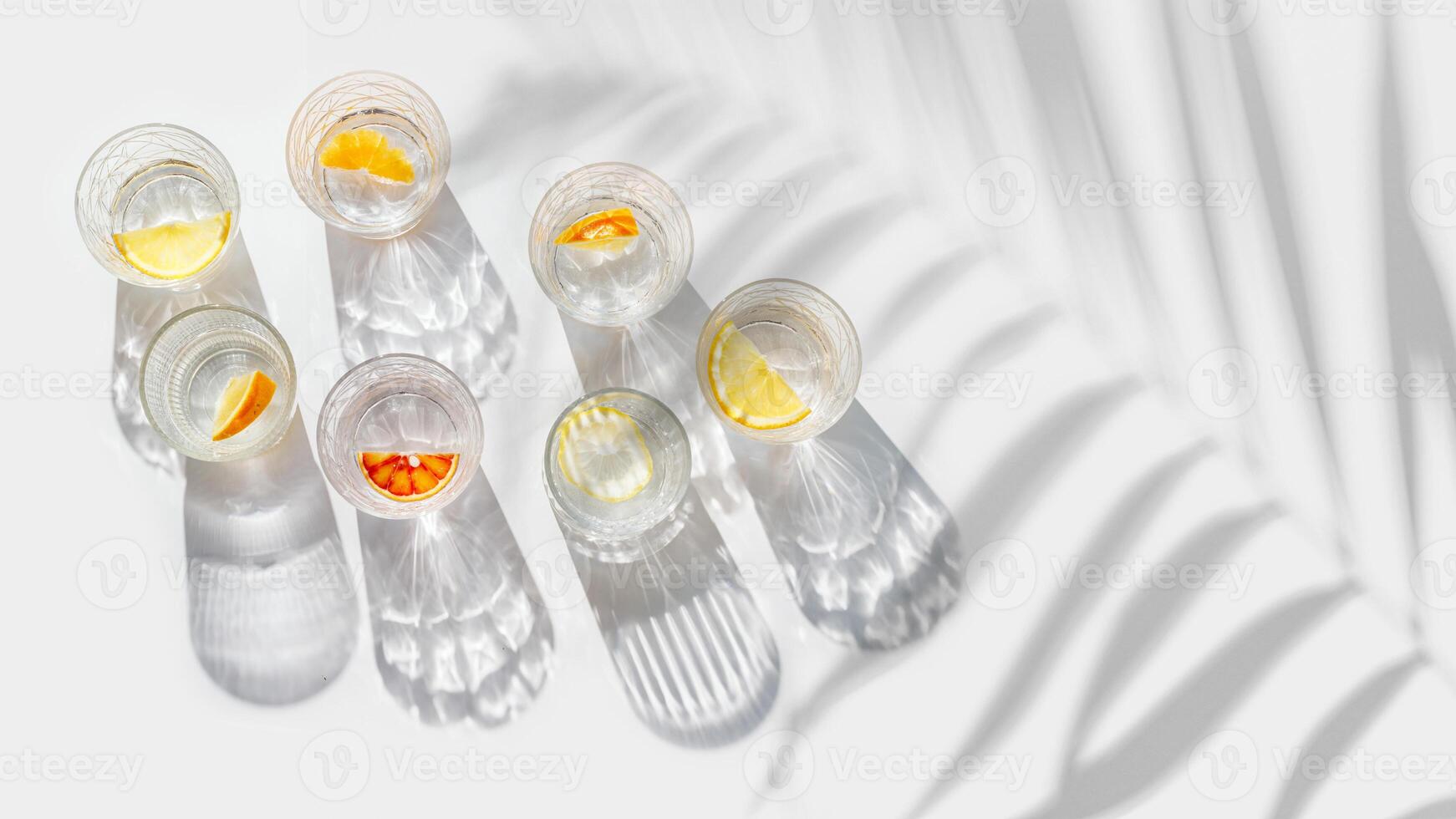 Crystal glasses with citrus slices in water on white background with palm leaf at sunlight, modern, minimal style banner with glass cups with water and citrus fruit, as lemon, orange photo