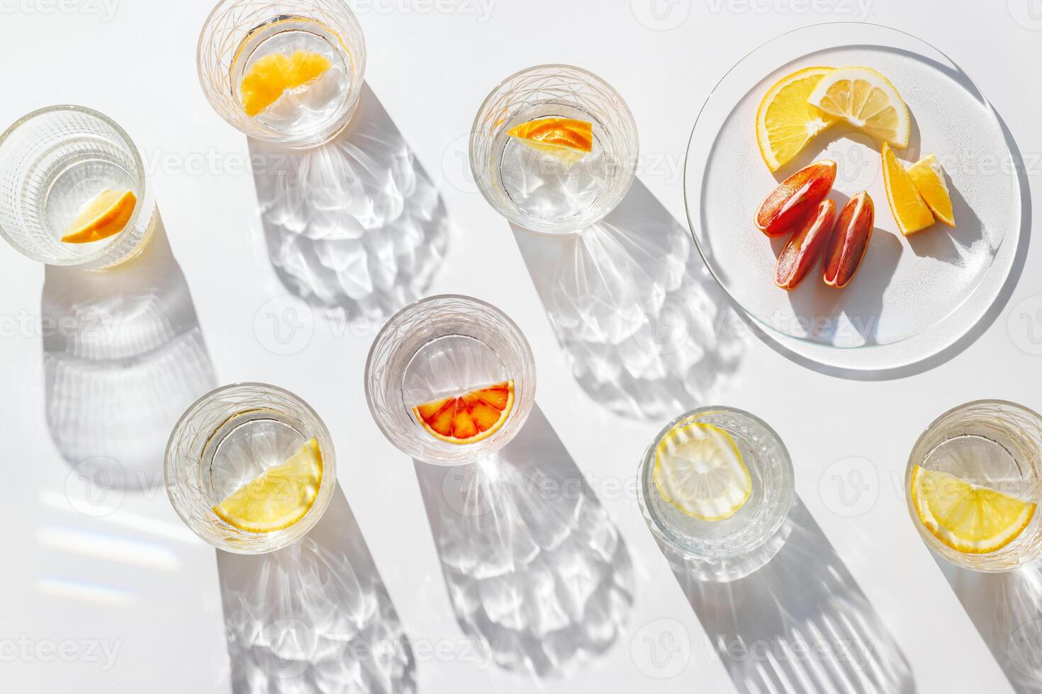 Fruit Infused Water in transparent Glasses with Shadows at sunlight on White background. Aesthetic view summer healthy drink with slices of lemon, orange with rainbow sun glare from glass cups, above. photo