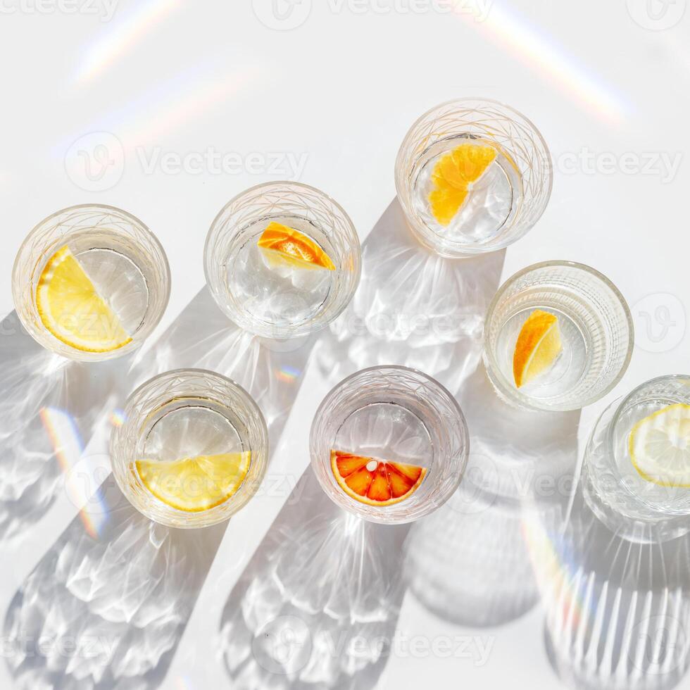 Fruit Infused Water in transparent Glasses with Shadows at sunlight on White background. Aesthetic view summer healthy drink with slices of lemon, orange with rainbow sun glare from glass cups photo