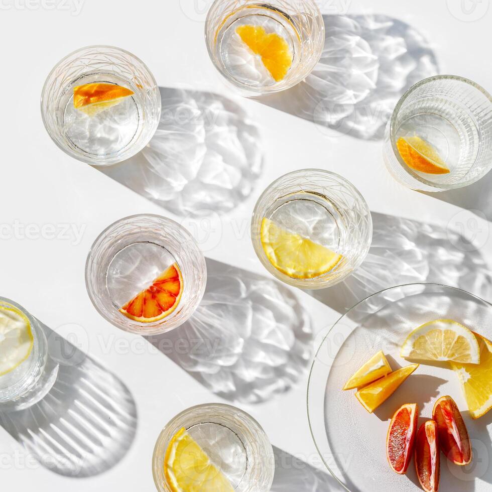 Citrus Infused Water in clear Glasses and plate, artistic light reflections and shadow in natural sunlight. Summer drink concept, minimal pattern, flavored water with yellow red slices of fruit, photo
