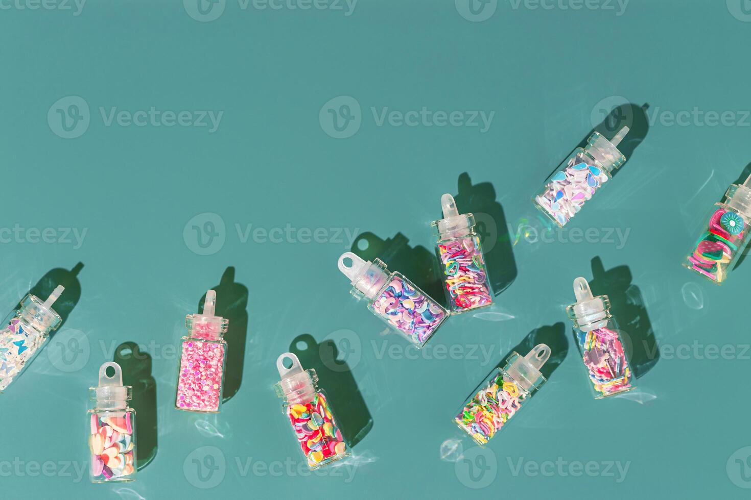 Assortment colored glitters for nail art and makeup in glass jars on teal background Trend Flat lay with glitter for nails in bottles with sahdows and glare, visual for manicure master photo
