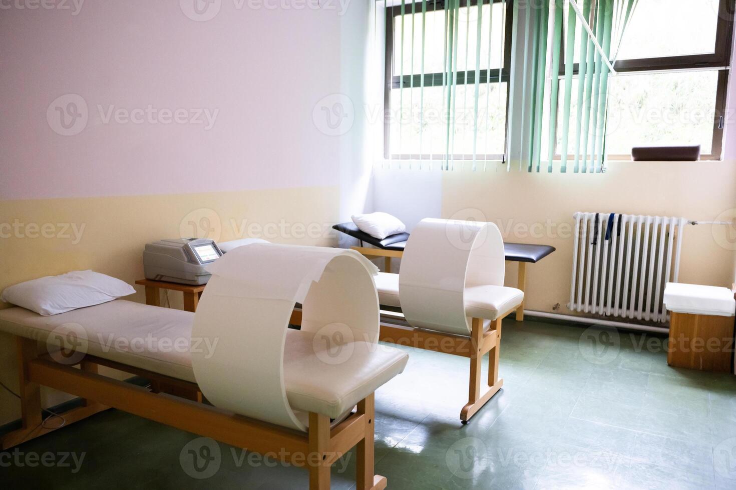 A well-lit physiotherapy treatment room featuring specialized medical equipment, therapy beds, and a comfortable clinical setting. The room has a clean and organized appearance with natural light from photo