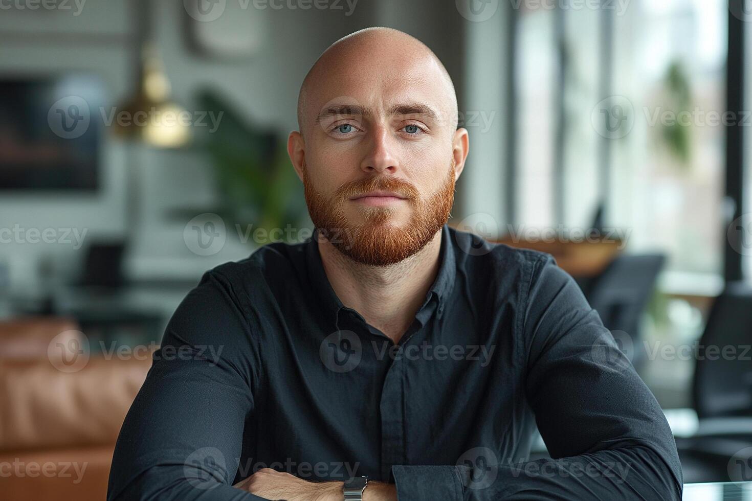 A young bald businessman with a red beard sits at a table in an office. Businessman is sitting in chair at his desk. photo