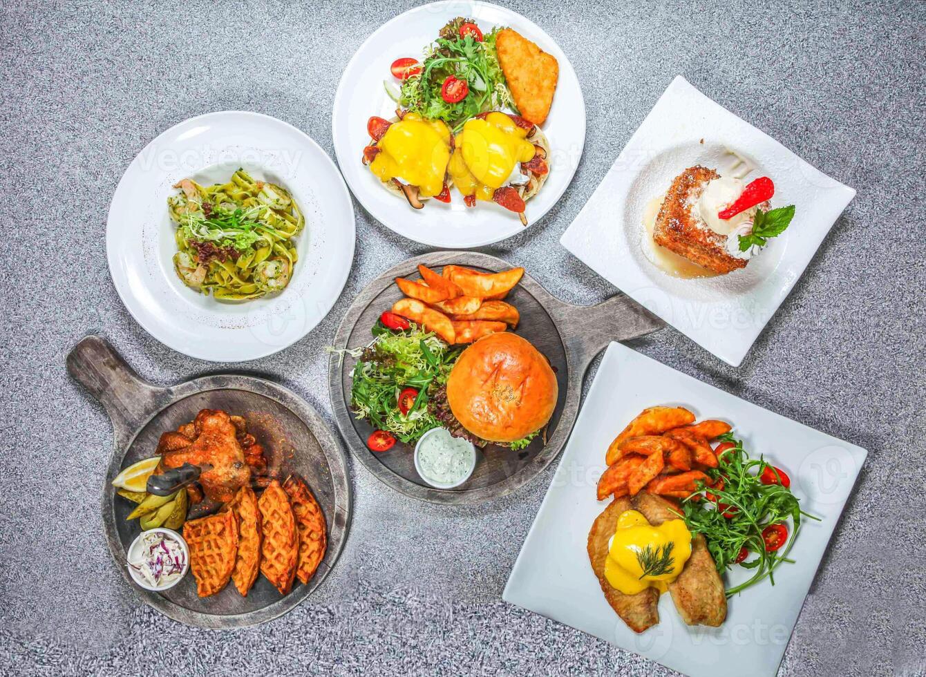Kiwi Lamb Burger, Chicken Wings and Waffles, Shrimp pesto sauce pasta, Fried French Toast Cube, Fried Fish Benedict, and Eggs Benedict served isolated on stone background top view of hong kong food photo