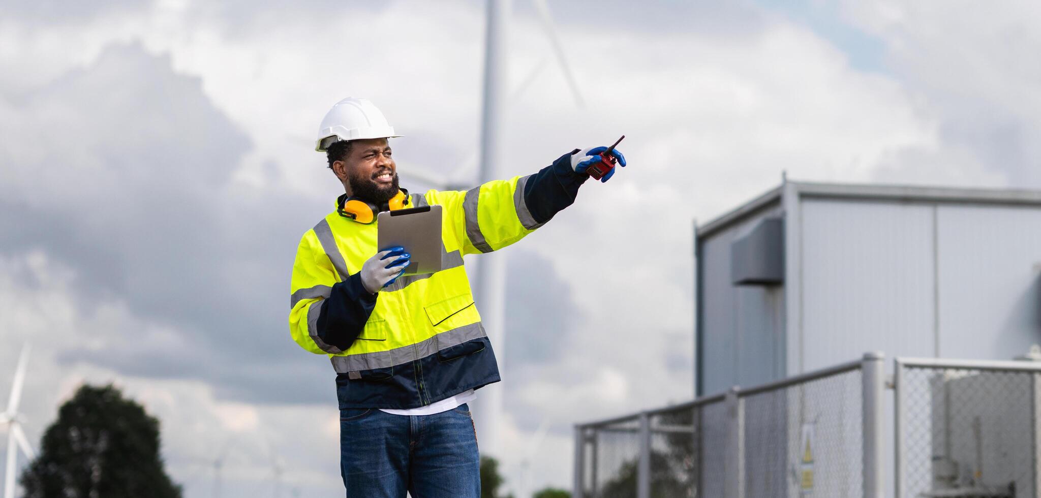 man in a yellow and black safety vest points to a wind turbine photo
