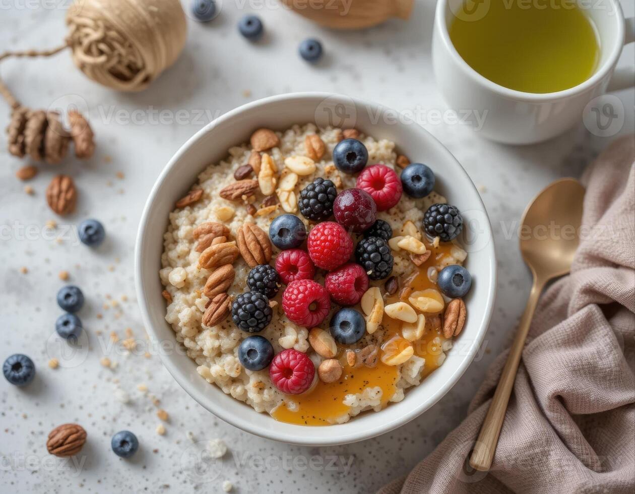On the light gray surface there is a plate with oatmeal, decorated with fresh berries ,raspberries, blueberries, blackberries, and nuts, sprinkled with honey. Next to a cup of green tea, top view photo