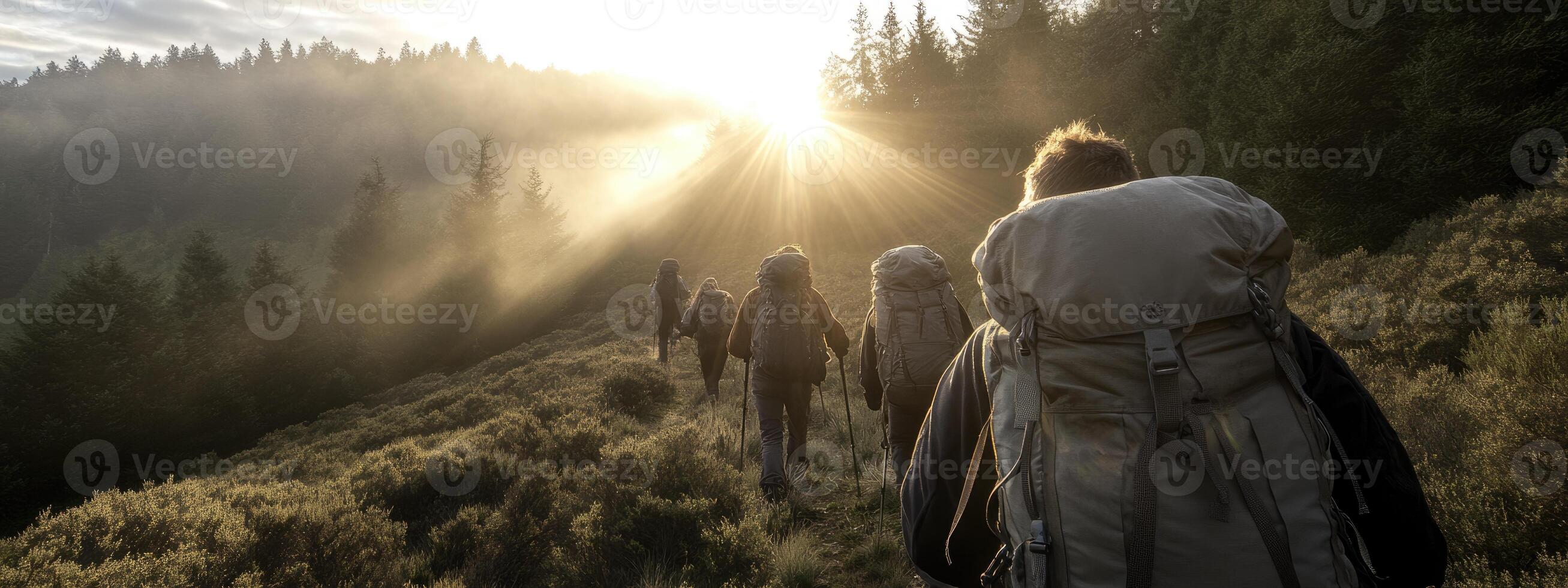 A group of hikers with large backpacks walks along a forested mountain trail during sunrise, with sunlight filtering through the trees and mist in the air photo