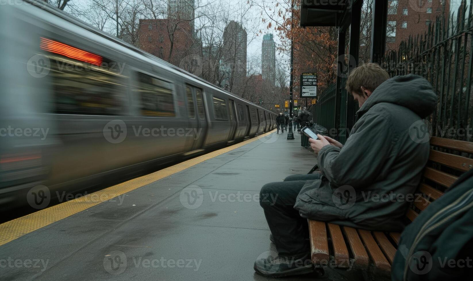 A man sits on a bench at a train station, focused on his phone as a subway train speeds past him. The atmosphere suggests a chilly day with a bustling city backdrop photo