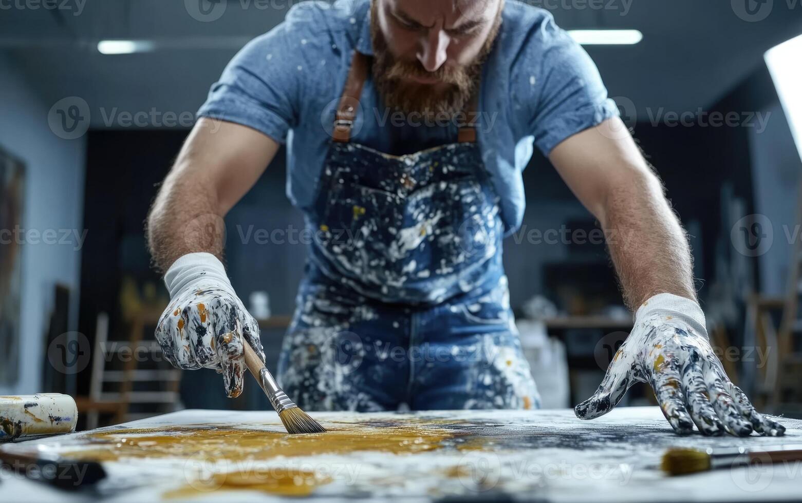 An artist is focused on his craft while mixing vibrant paint colors on a large canvas. He wears a paint-splattered apron and gloves, showing his dedication to his work in the studio photo