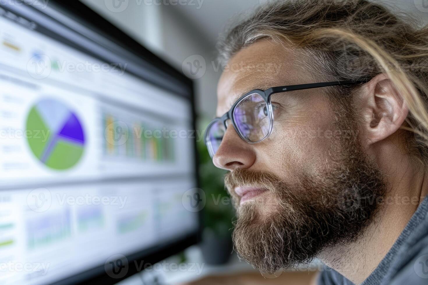 A man with long hair and glasses thoughtfully examines various data visualizations displayed on a large monitor in a contemporary office setting photo