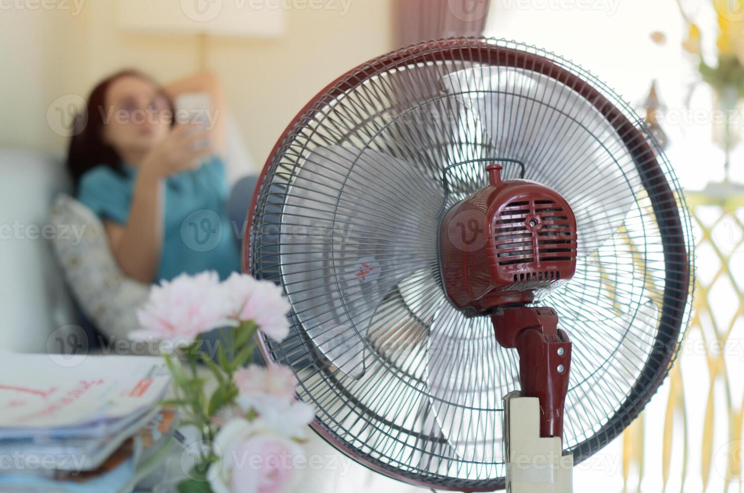 Asian woman sitting on sofa and holding her smart phone in front of electric fan at home. photo