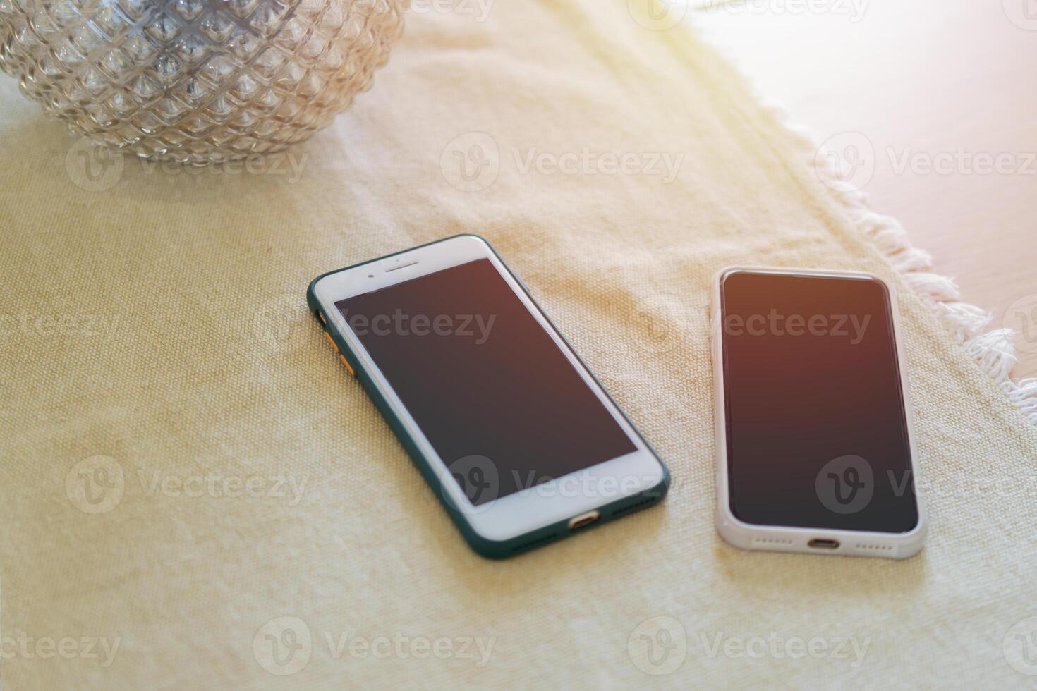 Two mobile phones on the dining room table with a tablecloth near by glass vase. photo