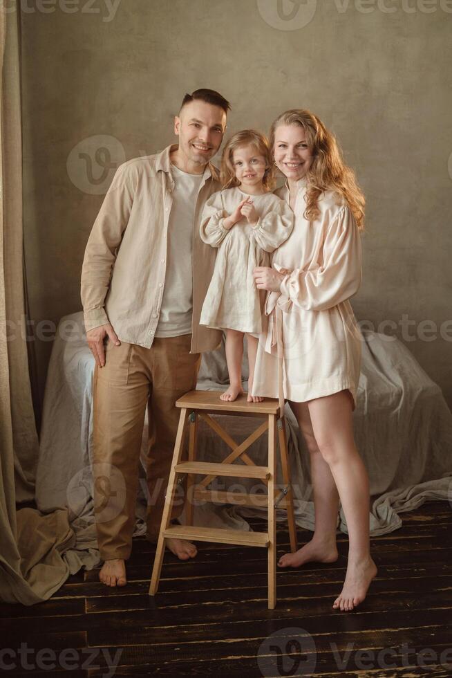Family portrait captured in a cozy indoor setting featuring a smiling father, mother, and daughter standing on a stool against a neutral background photo