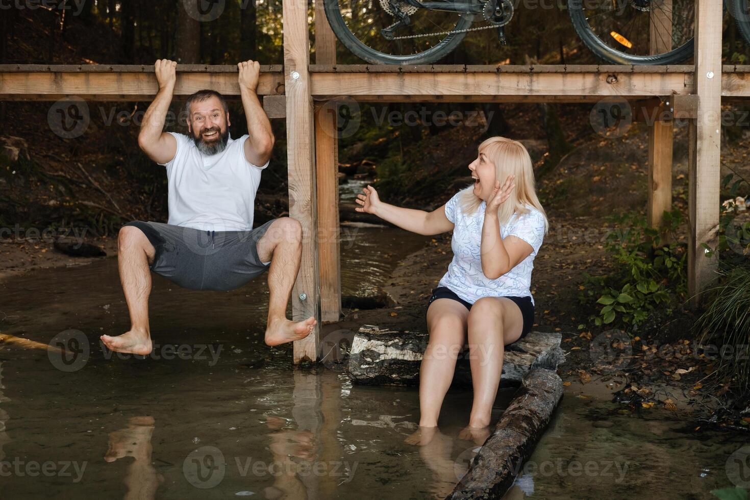 Friends enjoying a summer day by the water, one hanging from a platform and the other seated on a log, both laughing joyfully in a natural setting photo