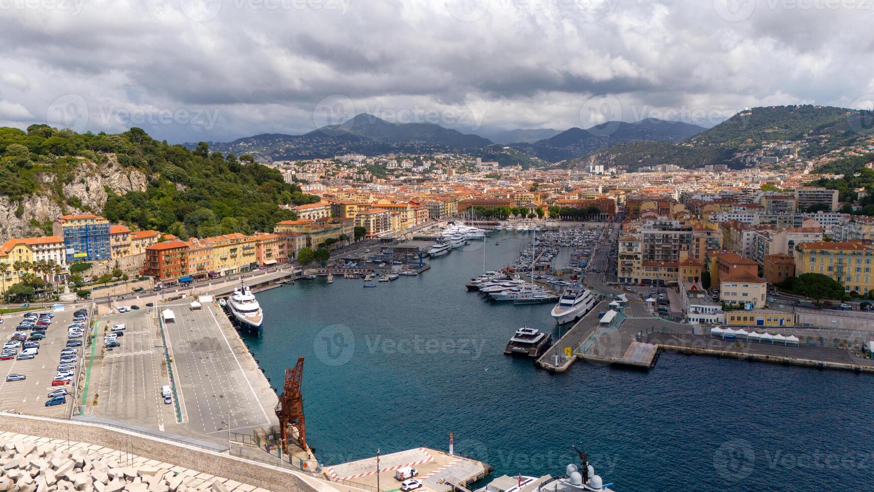A panoramic view of Nice Port in France, featuring yachts, a waterfront, and colorful buildings against green hills and a cloudy sky. photo