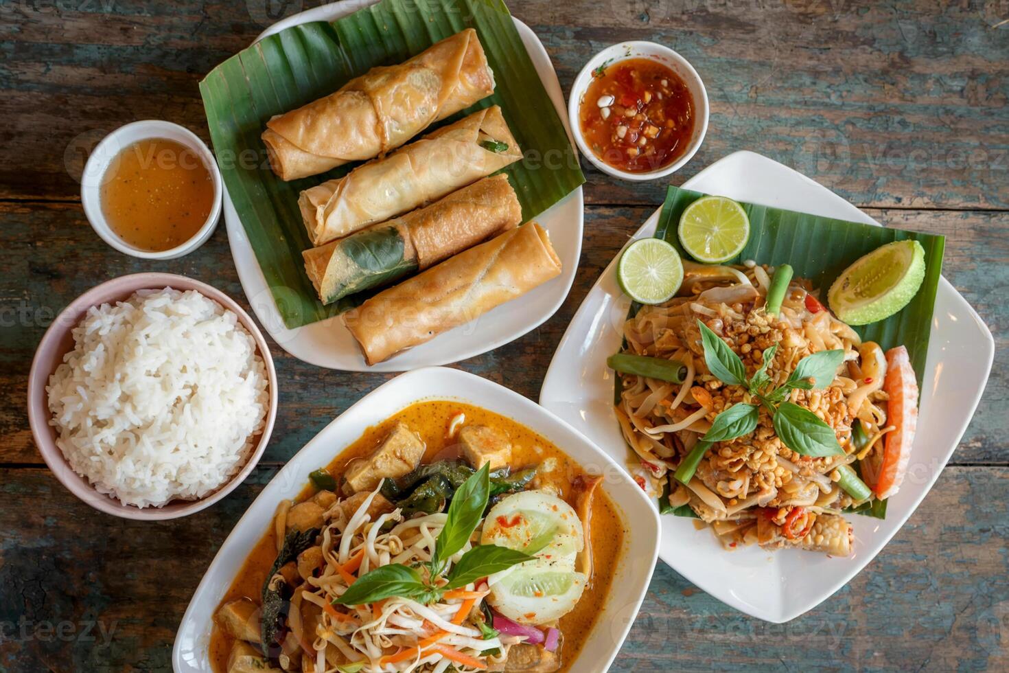 An assortment of vibrant Thai dishes, including spring rolls, rice, and a flavorful noodle dish, is beautifully arranged on a rustic wooden table. photo