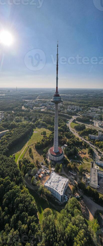 Tall telecommunications tower surrounded by greenery and urban landscape. Aerial view of a modern observation tower with a red and white antenna photo