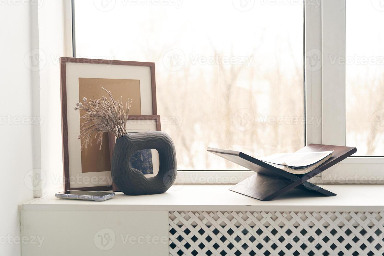 Minimalist composition on the windowsill near the window with a copy space. A vase with dried flowers, a book, and photo frames. Home Decor Template
