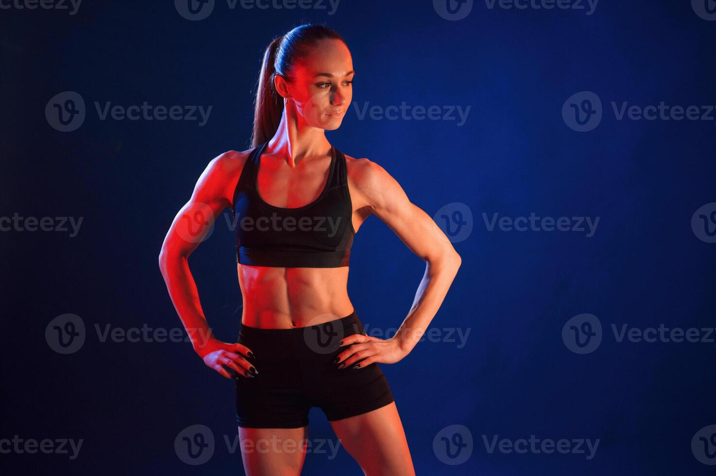Hands are on the waist, standing. Beautiful muscular woman is indoors in the studio with neon lighting photo