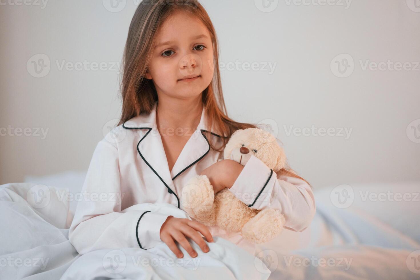 Little girl with teddy bear in hands is sitting on the bed at home photo