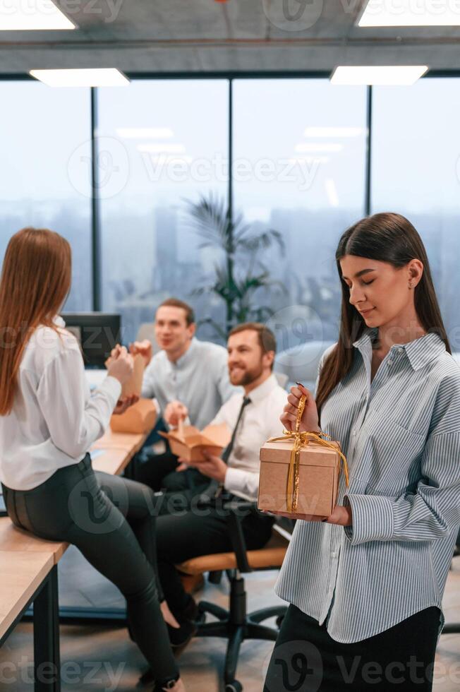 Woman is, standing and holding holidays gift. Group of coworkers are eating food from eco boxes in the office photo