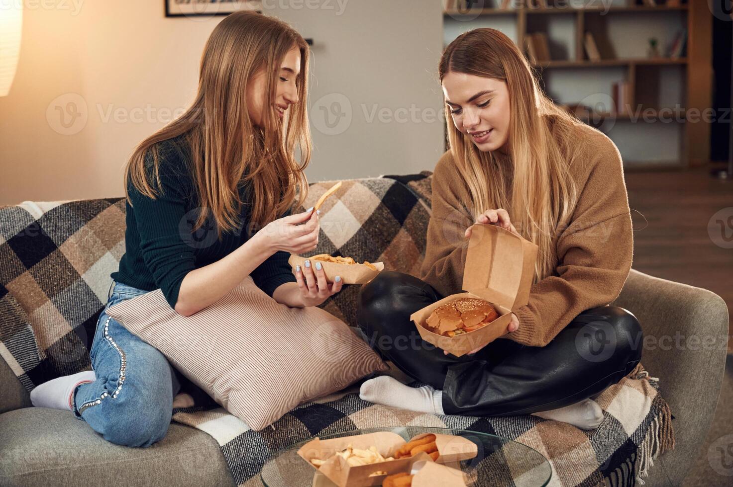 Smiling, enjoying the meal.. Two, women are sitting and eating burger, French fries and chicken nuggets indoors together photo