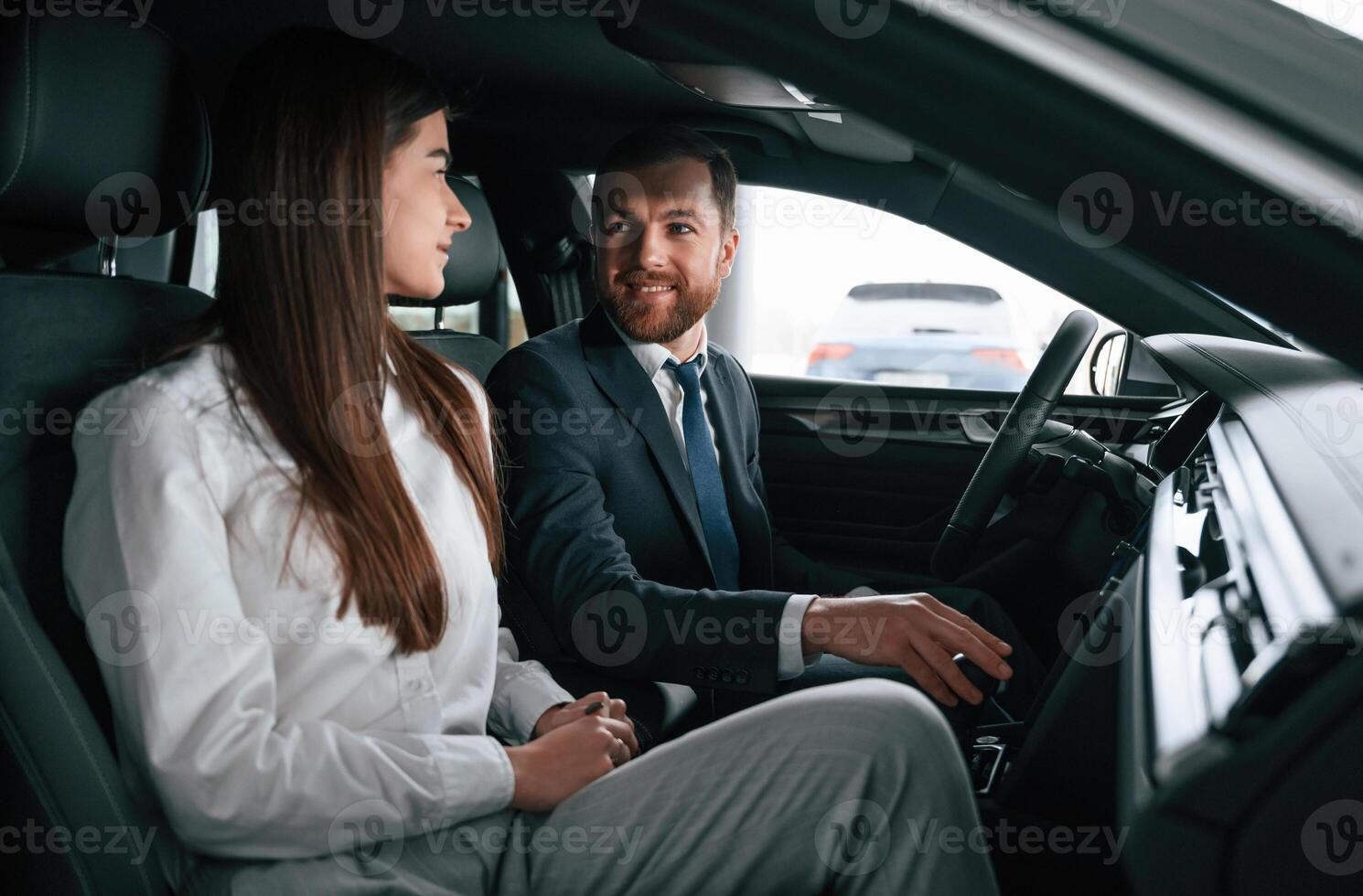 Man, in suit and tie is sitting with young woman, customer, in the automobile and showing the interior of the car photo