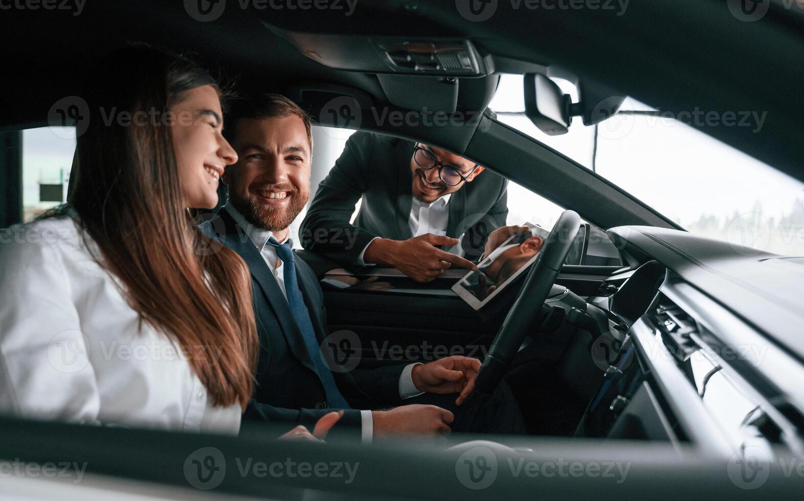 Side, view. Car showroom worker is consulting man and woman that are sitting inside of automobile photo