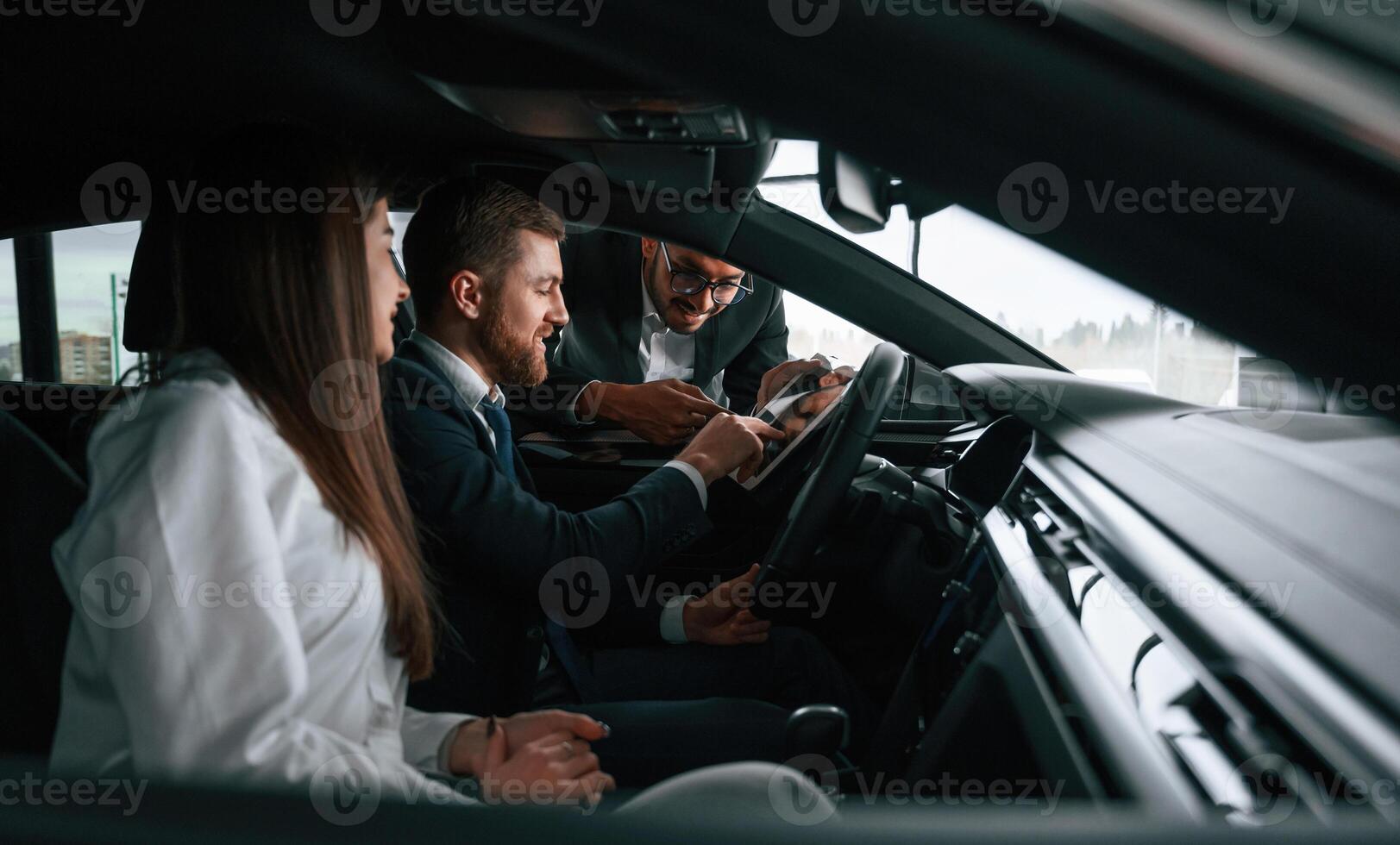 Side view. Car showroom, worker, is consulting man and woman that are sitting inside of automobile photo