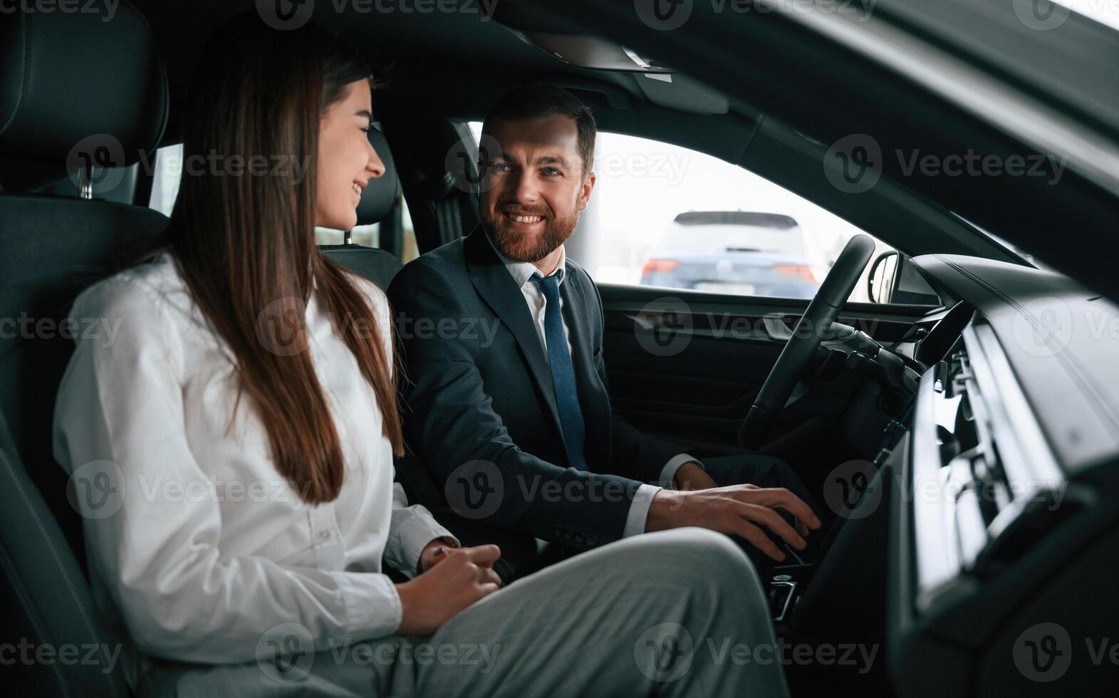 Man in, suit and tie is sitting with young woman, customer, in the automobile and showing the interior of the car photo