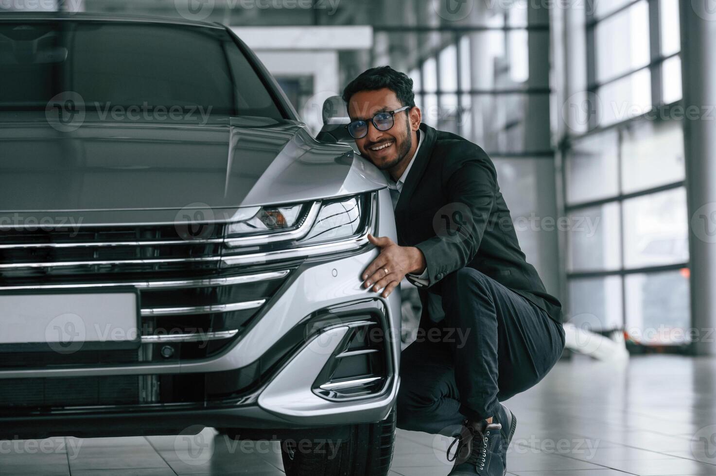 Embracing ,the transport. Handsome indian man in suit is in the car showroom with the vehicle indoors photo
