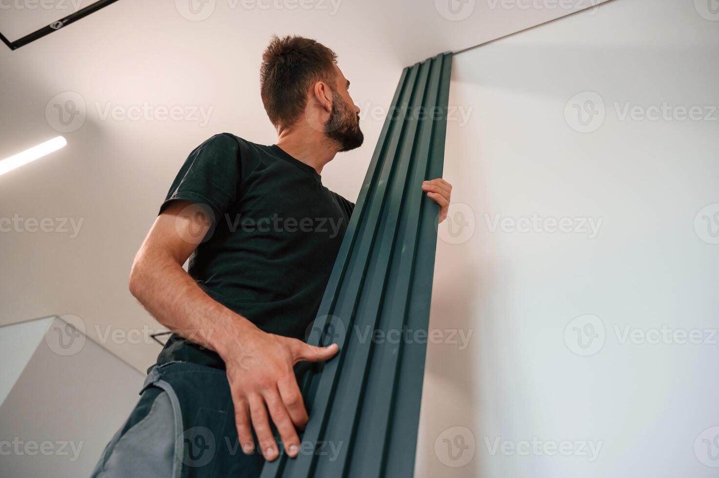 Holding tall ,heating radiator. Plumber worker is in empty room of a newly built house photo