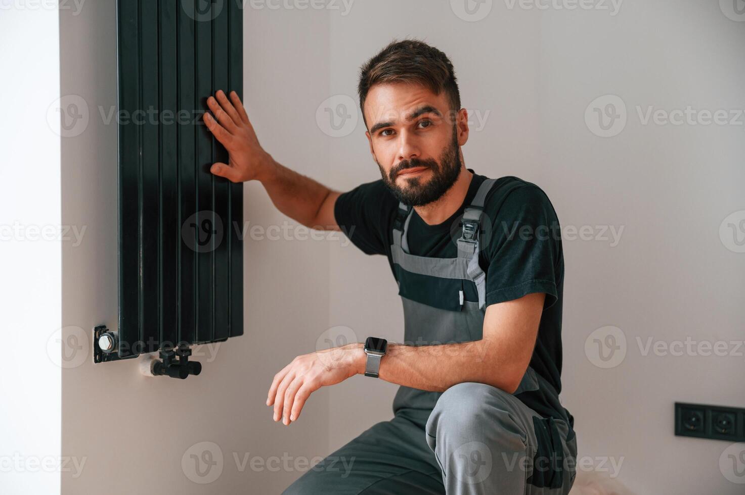 Looking at, the ,camera. Plumber worker installing heating radiator in empty room of a newly built house photo