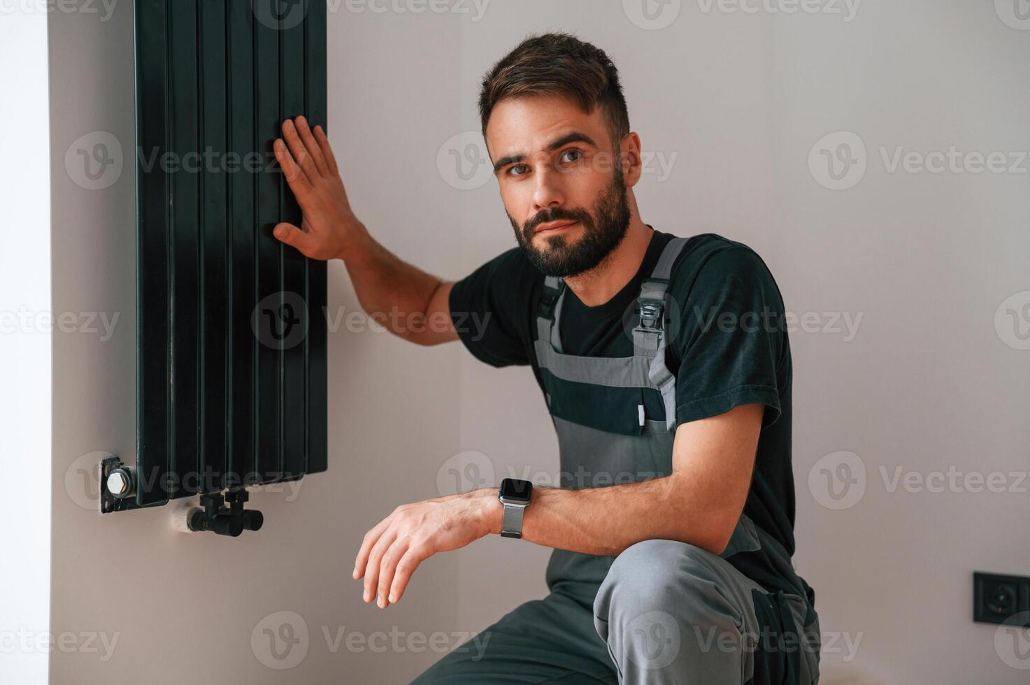 Looking at the camera. Plumber worker installing heating radiator in empty room of a newly built house photo