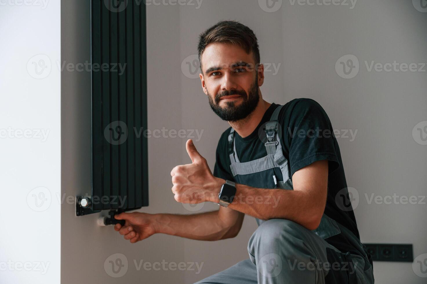 Sitting, and showing thumb, up. Plumber worker installing heating radiator in empty room of a newly built house photo