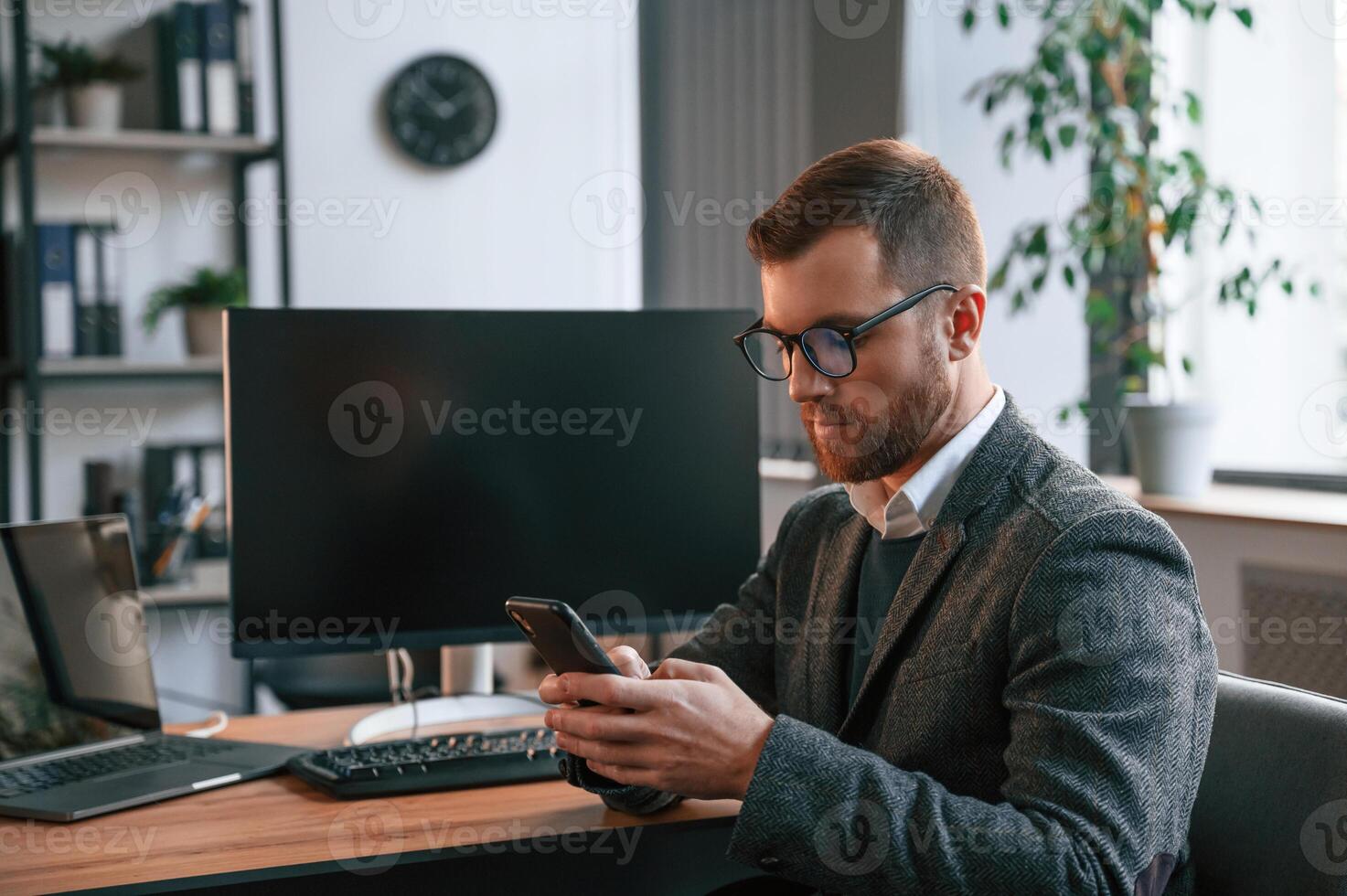 Sitting, using smartphone. Businessman in suit is in the office with computer photo