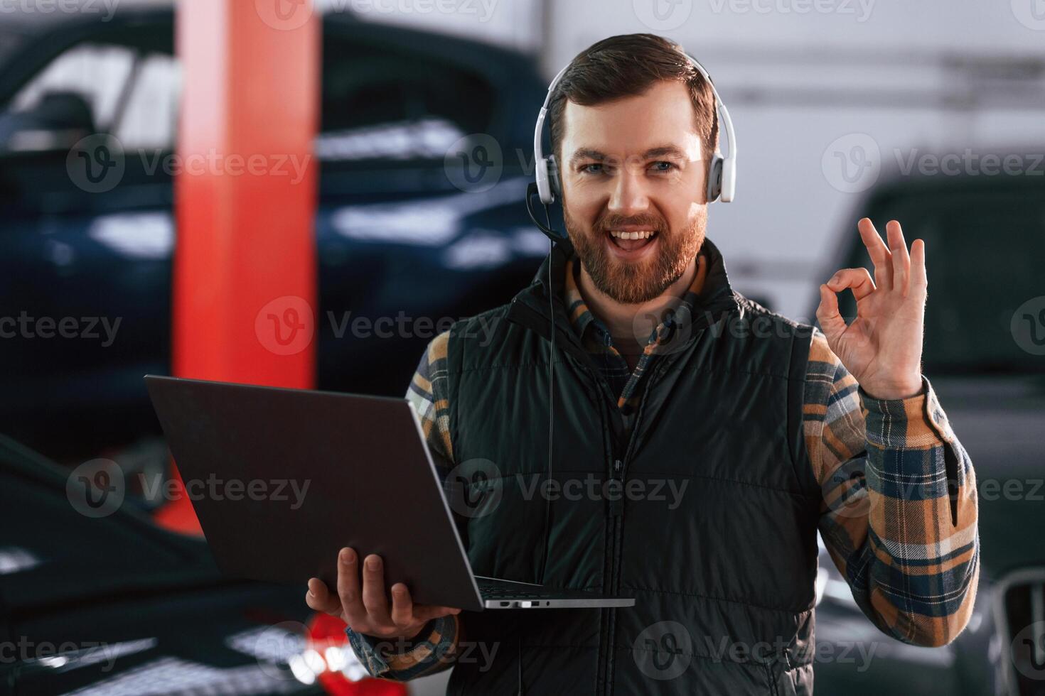 Smiling, standing, holding laptop. In headphones. Man in uniform is working in the auto salon photo