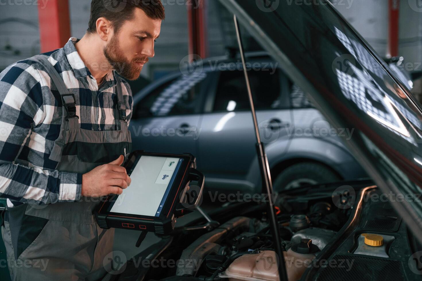 Standing near, the car with hood opened. Holding tablet. Man in uniform is working in the auto salon photo